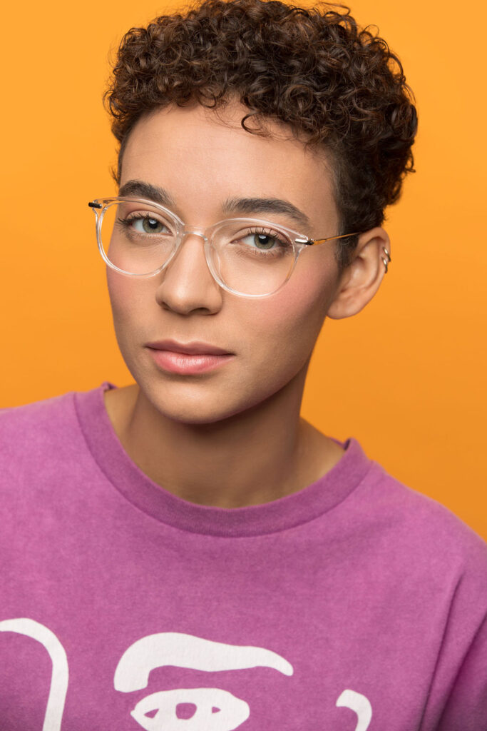 Headshot of Aislinn Brophy, a mixed-race Black and white person with short curly brown hair. They wear clear glasses and a brightly colored purple shirt with an abstract print of an eye.