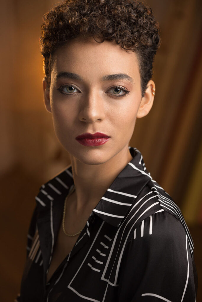 Headshot of Aislinn Brophy, a mixed-race Black and white person with short curly brown hair. The photo is lit dramatically, and Aislinn wears dark makeup with bright red lipstick.