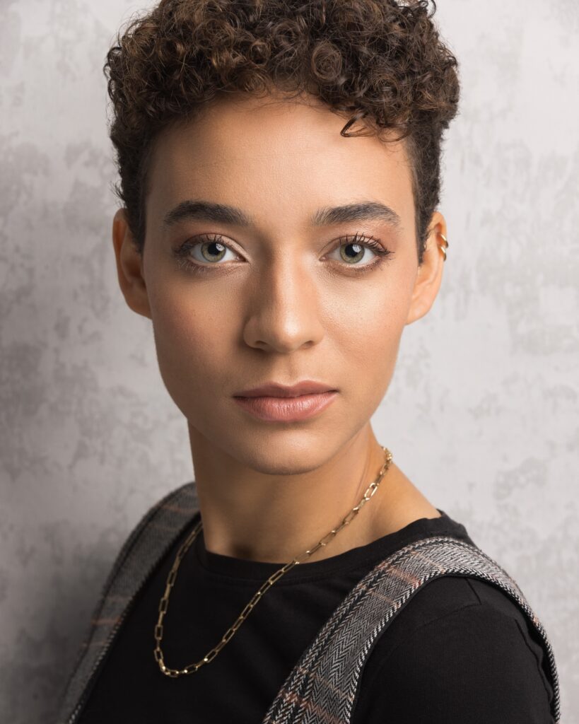 Headshot of Aislinn Brophy, a mixed-race Black and white person with short curly brown hair. They face sideways at the camera with an open, thoughtful expression.
